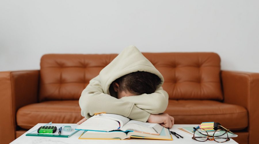 child in beige hoodie leaning forward on table feeling exhausted while studying