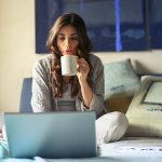 woman in grey jacket sits on bed uses grey laptop