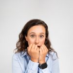 A brunette woman wearing a nice dress shirt and smart watch, wide-eyed and holding her closed hands close to her mouth in anxiety.