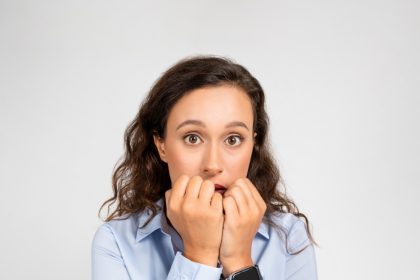 A brunette woman wearing a nice dress shirt and smart watch, wide-eyed and holding her closed hands close to her mouth in anxiety.