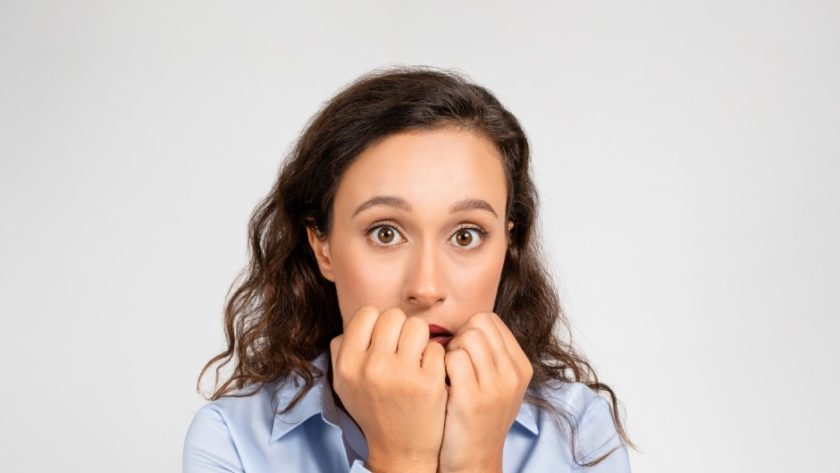 A brunette woman wearing a nice dress shirt and smart watch, wide-eyed and holding her closed hands close to her mouth in anxiety.