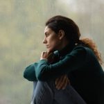 A young woman on a rainy day looking sad while she sits with her knees curled up and looks out the window.