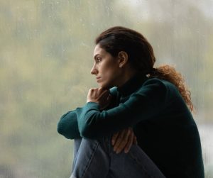 A young woman on a rainy day looking sad while she sits with her knees curled up and looks out the window.