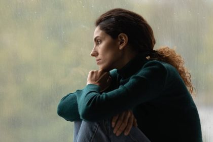 A young woman on a rainy day looking sad while she sits with her knees curled up and looks out the window.