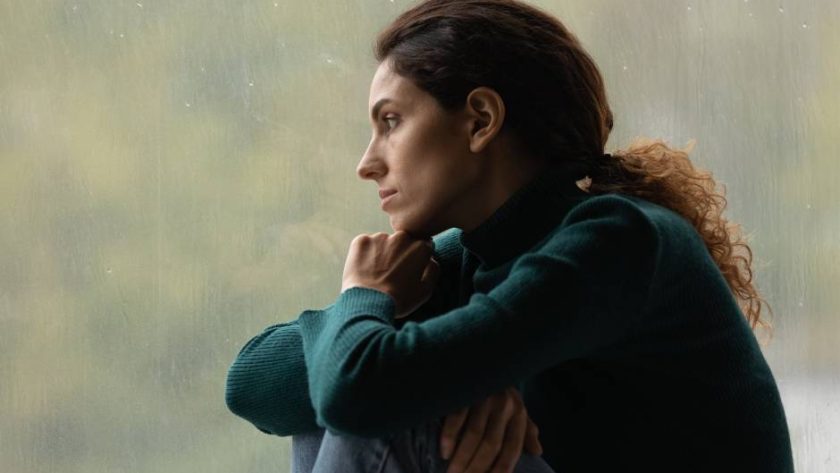 A young woman on a rainy day looking sad while she sits with her knees curled up and looks out the window.