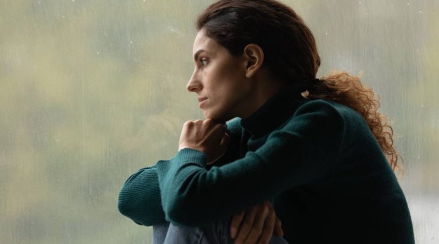 A young woman on a rainy day looking sad while she sits with her knees curled up and looks out the window.