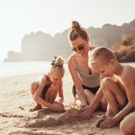 A mother and her young son and daughter building sandcastles on the beach. Everyone is wearing a bathing suit and smiling.