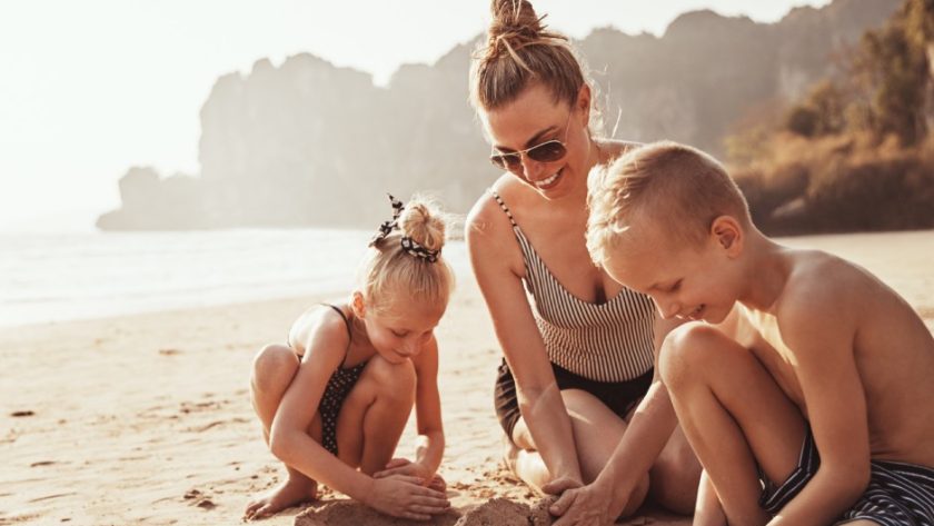 A mother and her young son and daughter building sandcastles on the beach. Everyone is wearing a bathing suit and smiling.