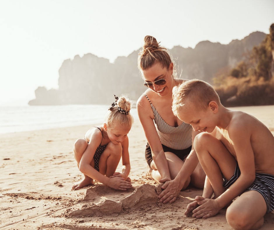 A mother and her young son and daughter building sandcastles on the beach. Everyone is wearing a bathing suit and smiling.