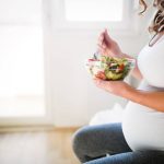 A pregnant woman in a white tank top sitting down as she eats a colorful salad full of nutrients.