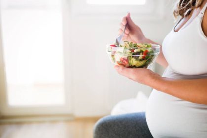 A pregnant woman in a white tank top sitting down as she eats a colorful salad full of nutrients.