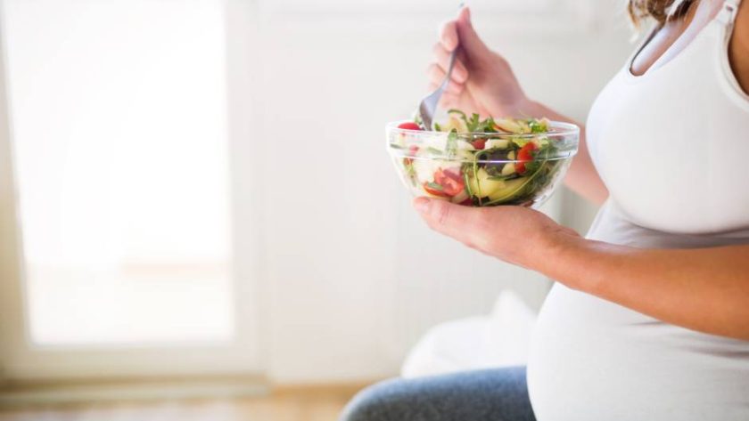 A pregnant woman in a white tank top sitting down as she eats a colorful salad full of nutrients.