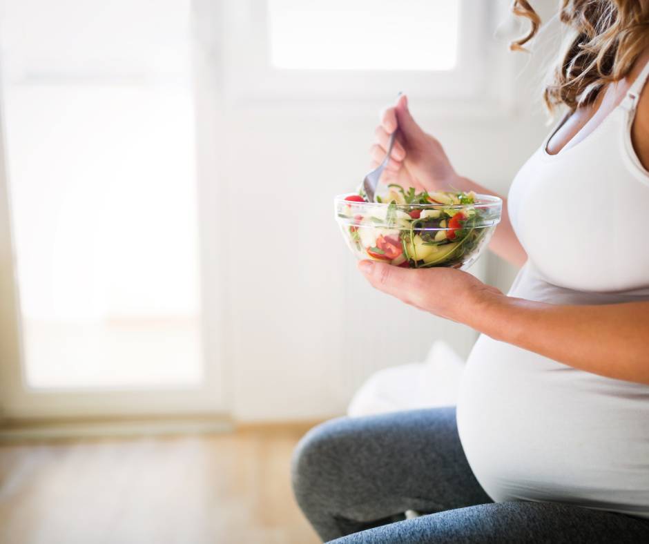 A pregnant woman in a white tank top sitting down as she eats a colorful salad full of nutrients.