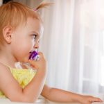 A toddler girl wearing a yellow dress and a ponytail, while using a purple pacifier despite being older than 12 months.