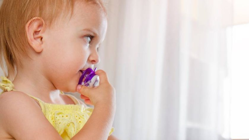 A toddler girl wearing a yellow dress and a ponytail, while using a purple pacifier despite being older than 12 months.