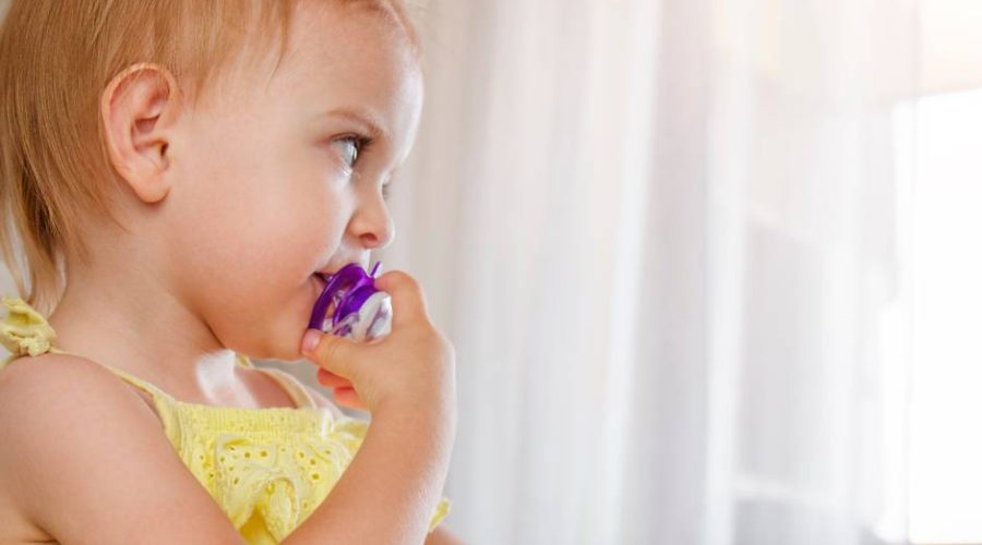 A toddler girl wearing a yellow dress and a ponytail, while using a purple pacifier despite being older than 12 months.