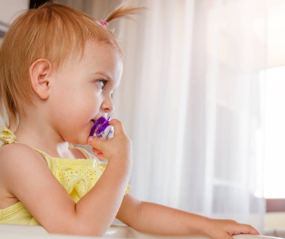 A toddler girl wearing a yellow dress and a ponytail, while using a purple pacifier despite being older than 12 months.