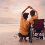 A parent with an older child in a wheelchair enjoying the sunset together on a beach and holding hands.