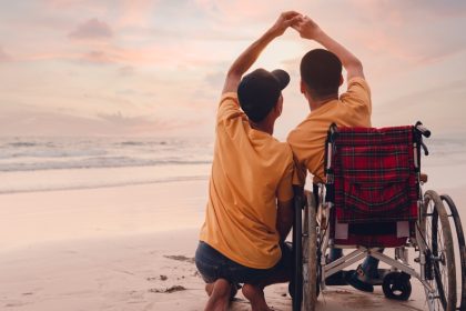 A parent with an older child in a wheelchair enjoying the sunset together on a beach and holding hands.