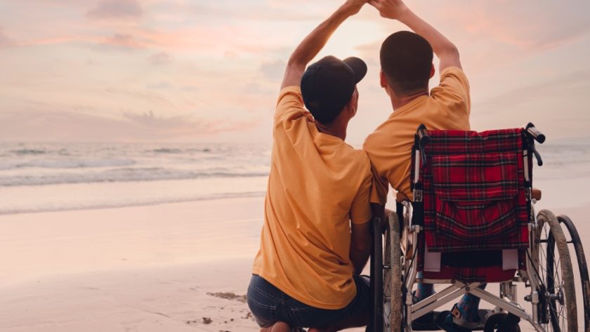 A parent with an older child in a wheelchair enjoying the sunset together on a beach and holding hands.