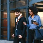 Two well-dressed office workers leaving a building. One of them carries a tablet and the other carries a laptop bag.