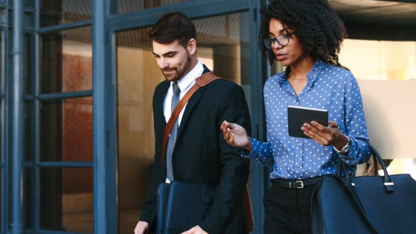 Two well-dressed office workers leaving a building. One of them carries a tablet and the other carries a laptop bag.