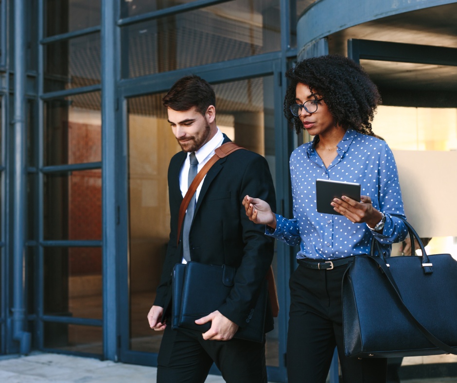 Two well-dressed office workers leaving a building. One of them carries a tablet and the other carries a laptop bag.