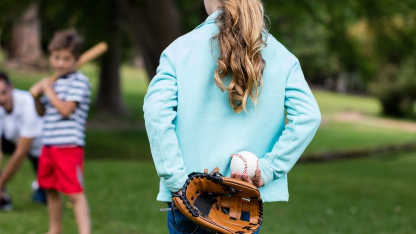 A girl standing in a park with a baseball mitt in her hand. She is preparing to toss the ball to her family.