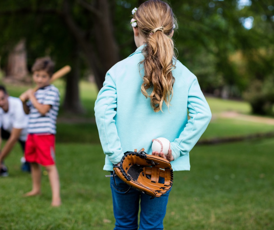 A girl standing in a park with a baseball mitt in her hand. She is preparing to toss the ball to her family.