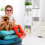 A toddler sitting on two luggage bags, ready to go on vacation. She’s wearing a sun hat and glasses, hugging a stuffed bear.