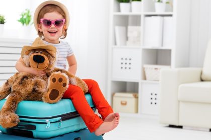 A toddler sitting on two luggage bags, ready to go on vacation. She’s wearing a sun hat and glasses, hugging a stuffed bear.