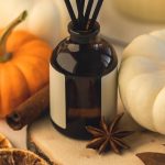 A glass bottle diffuser sits on a table next to a white pumpkin, an orange pumpkin, a clove, and a cinnamon stick.