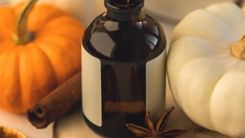 A glass bottle diffuser sits on a table next to a white pumpkin, an orange pumpkin, a clove, and a cinnamon stick.