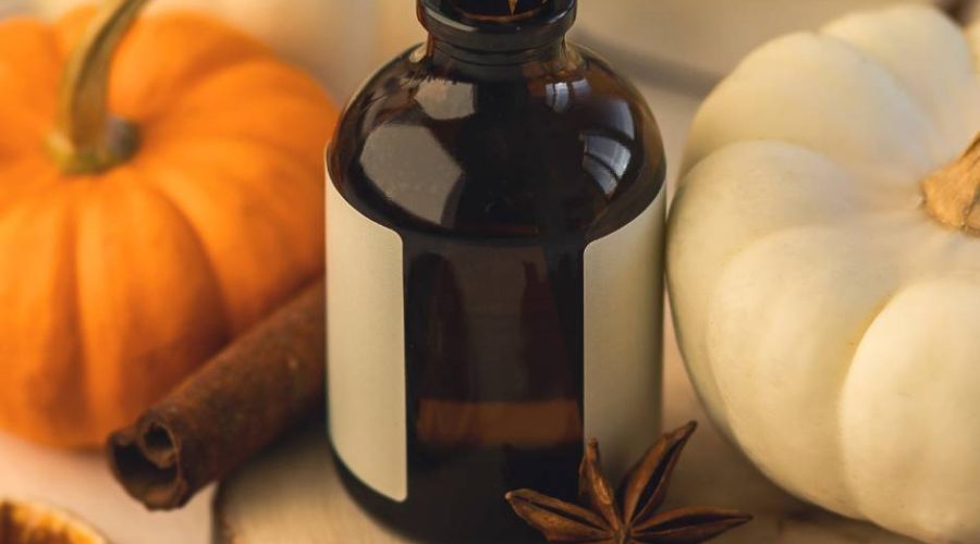 A glass bottle diffuser sits on a table next to a white pumpkin, an orange pumpkin, a clove, and a cinnamon stick.