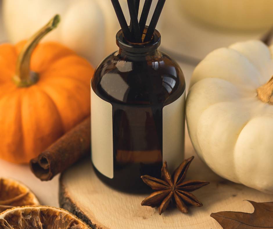 A glass bottle diffuser sits on a table next to a white pumpkin, an orange pumpkin, a clove, and a cinnamon stick.