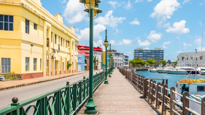 A wooden walkway with a rope barrier near a body of water with boats and a row of buildings painted in different colors.