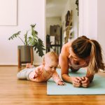 A woman is wearing workout clothes and doing a plank on a yoga mat at home. A young baby is laying on her tummy next to her.