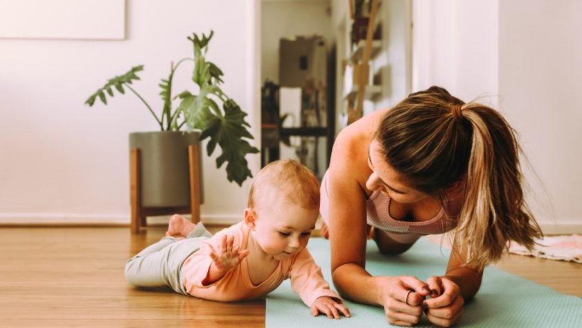 A woman is wearing workout clothes and doing a plank on a yoga mat at home. A young baby is laying on her tummy next to her.