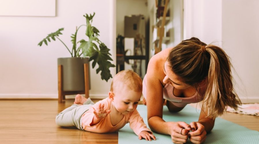 A woman is wearing workout clothes and doing a plank on a yoga mat at home. A young baby is laying on her tummy next to her.