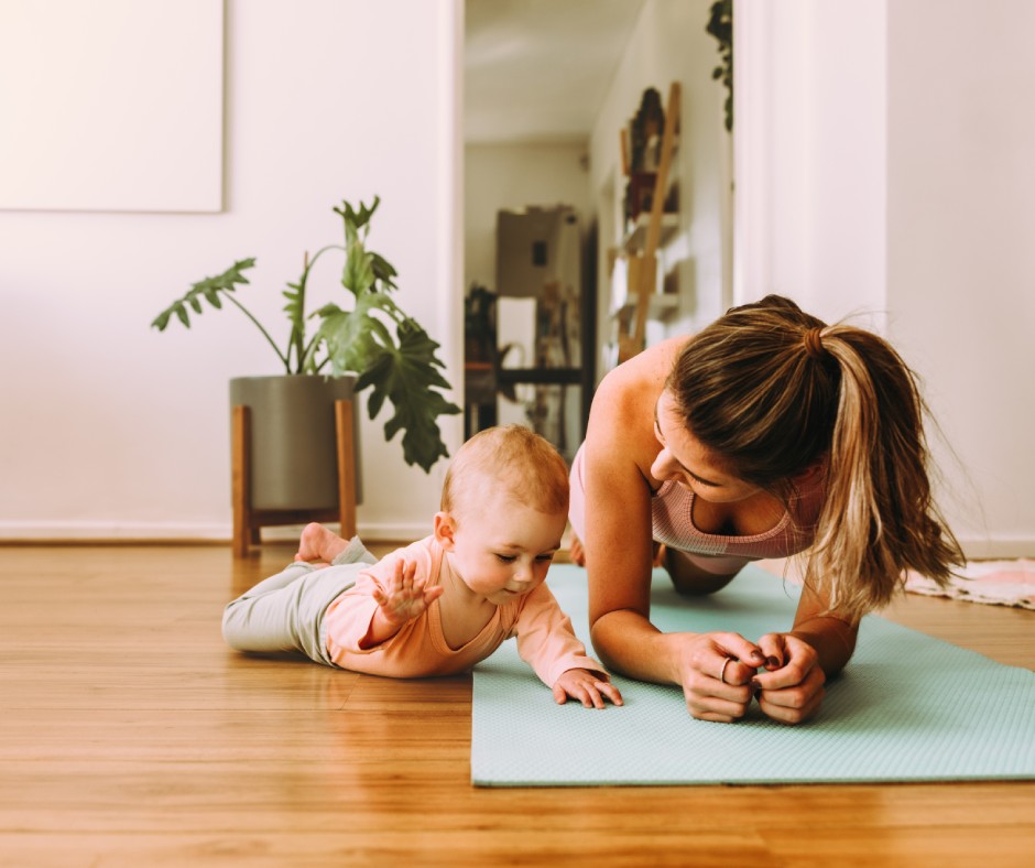 A woman is wearing workout clothes and doing a plank on a yoga mat at home. A young baby is laying on her tummy next to her.