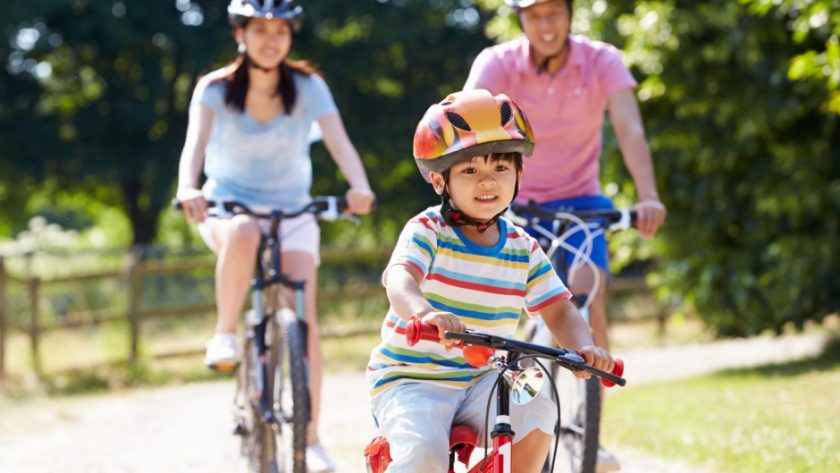 A family consisting of a mom, dad, and little boy riding bikes together down a path. They each wear a helmet.