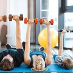 A family, consisting of the father, mother, and child, exercise in their living room by lifting dumbbells over their bodies.