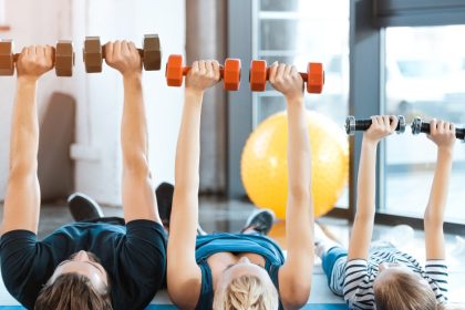 A family, consisting of the father, mother, and child, exercise in their living room by lifting dumbbells over their bodies.