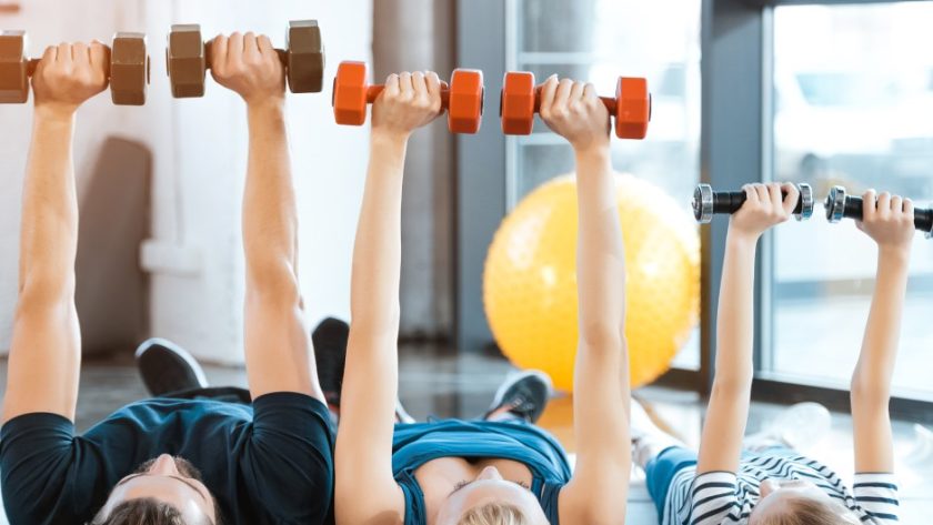 A family, consisting of the father, mother, and child, exercise in their living room by lifting dumbbells over their bodies.