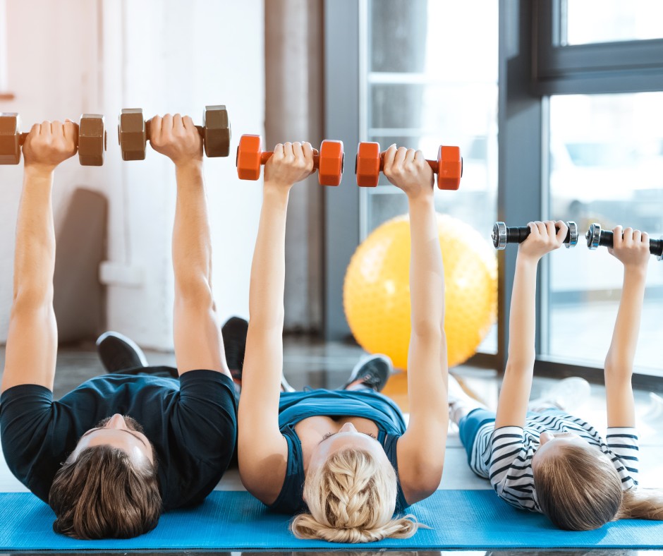 A family, consisting of the father, mother, and child, exercise in their living room by lifting dumbbells over their bodies.