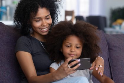 A mother and her daughter sitting on a couch. The mother is holding a smartphone and they're both looking at the screen.