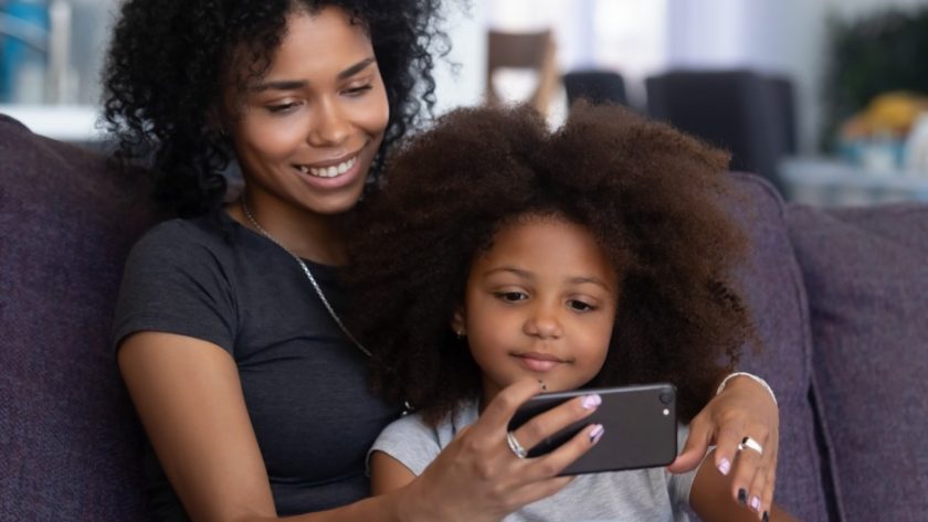 A mother and her daughter sitting on a couch. The mother is holding a smartphone and they're both looking at the screen.