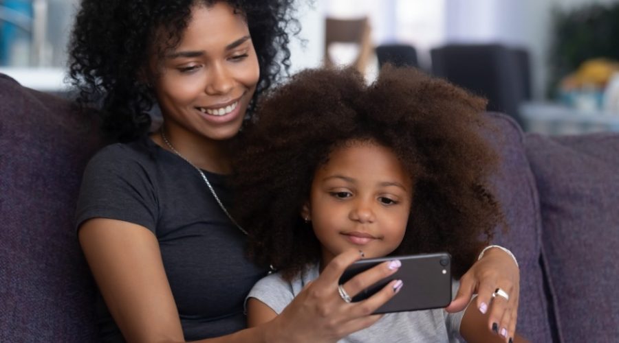 A mother and her daughter sitting on a couch. The mother is holding a smartphone and they're both looking at the screen.