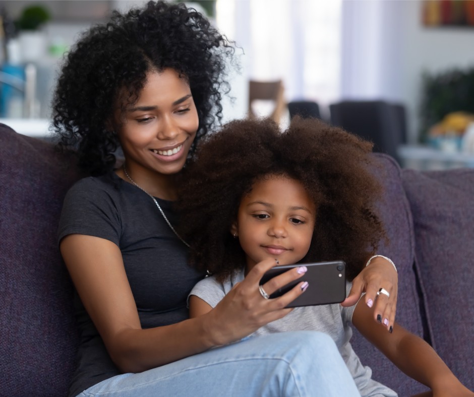 A mother and her daughter sitting on a couch. The mother is holding a smartphone and they're both looking at the screen.