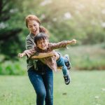 A mom holds her son off the ground, pretending he's an airplane. They are playing outside, surrounded by trees.